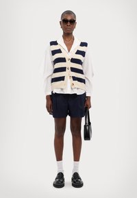 Kootud vest tumehallide ja kreemjate triipudega, nööpidega kinnitatud, kantud valge sordiini kohal, koos tumedate lühikeste pükste ja läikivate mustade kingadega. Mustad prillid.