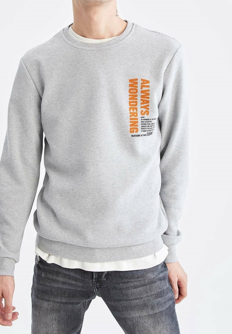 Homme portant un sweat-shirt texturé gris clair avec un texte orange "Always Wondering" et un petit texte noir sur la poitrine, associé à un jean foncé.