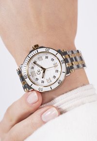 Srebrni i zlatni sat s bijelim teksturiranim brojčanikom, rimskim brojevima, kristalnim markerima i metalnom narukvicom s naizmjeničnim karikama.