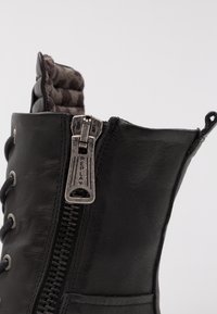 Zbliżenie czarnej skórzanej botki z metalowym zamkiem błyskawicznym oznaczonym "REPLAY" oraz sznurowadłami, ukazujące wyściełane wnętrze i pętelkę na pięcie.