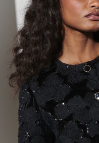 Prenda negra con textura y patrón floral, con lentejuelas y un adorno de piedra azul decorativa. Primer plano del escote y el área del hombro.