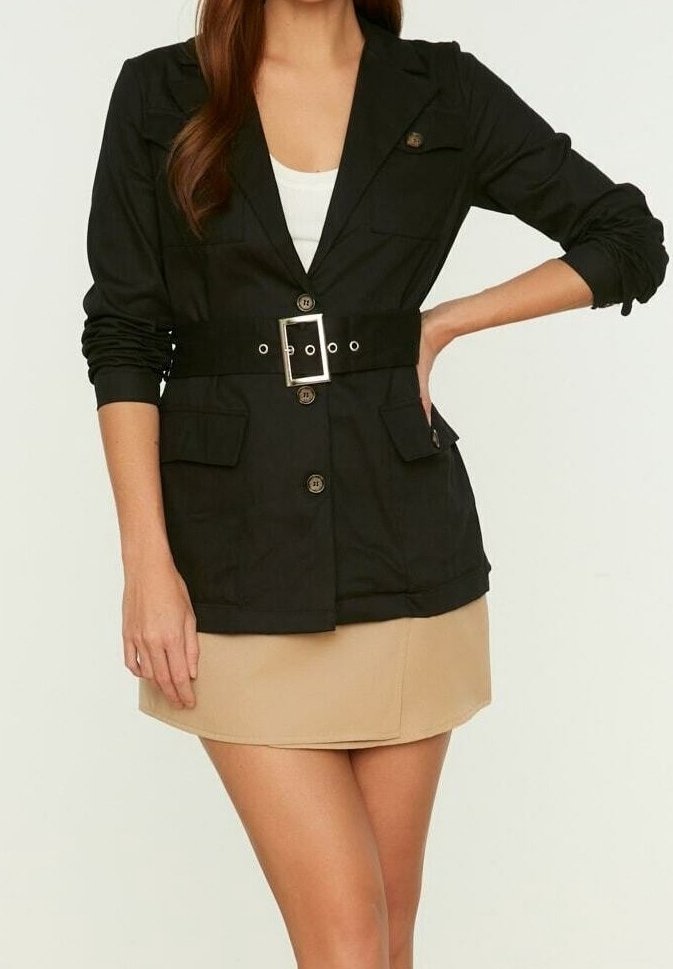 Blazer noir ceinturé avec col cranté, deux poches avant, fermeture à boutons et manches retroussées, porté sur une jupe beige.