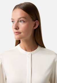 Witte blouse met lange mouwen, een ronde halslijn en een enkele knoopsluiting. Soepele stof met een subtiele glans. Geen patronen of versieringen.