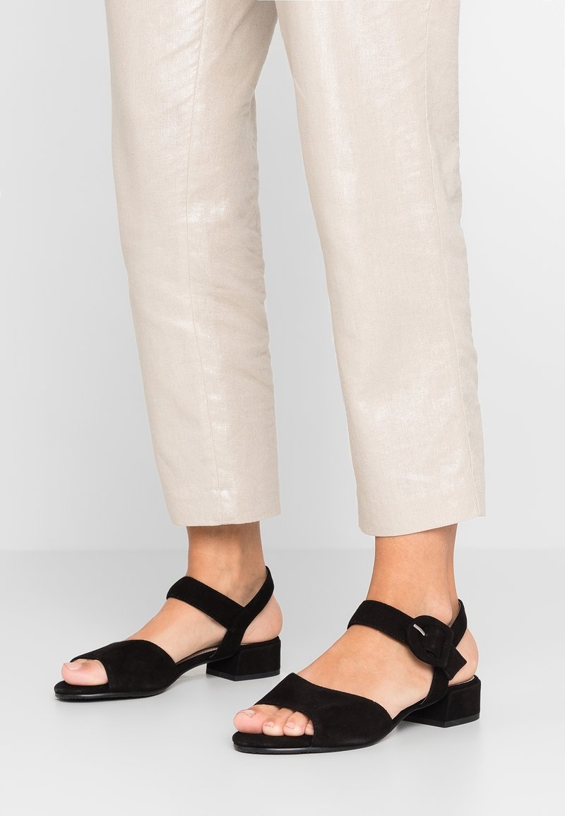 Jambes inférieures portant un pantalon beige court et des sandales noires à bout ouvert avec des talons carrés bas et des brides de cheville à boucle.