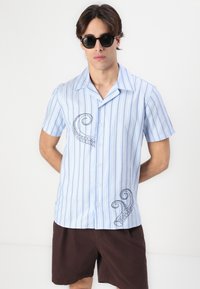 Kurzarm-Hemd in hellblau mit Streifen, Knopfverschluss und marineblauem Oktopus-Stickerei auf der linken Vorderseite. Das Material ist leicht und glatt.