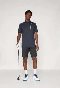 Muž v námorníckom golfovom polo tričku, čiernych šortkách, bielej golfovej rukavici a teniskách, drží golfovú palicu a stojí na jednoduchom pozadí.