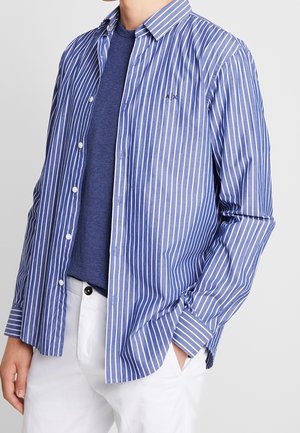 Homme portant une chemise à rayures verticales bleues et blanches boutonnée sur un t-shirt bleu à col rond et un pantalon blanc, main dans la poche.