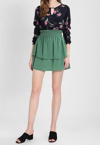 Granatowa bluzka w kwiatowy wzór z dekoltem na zapięcie, zestawiona z warstwową zieloną spódnicą. Stylizację dopełniają czarne mule z otwartymi palcami.