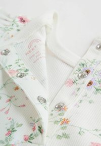 Mono con estampado floral en blanco, con textura acanalada, botones de presión y un cuello suave. Detalles en flores rosas, moradas y verdes.
