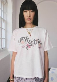 Moteris su tiesiais juodais plaukais, vilkinti balta T-shirt su katės iliustracijomis ir užrašu "katė Kristin", bei sluoksniuotais auksiniais kaklo papuošalais.