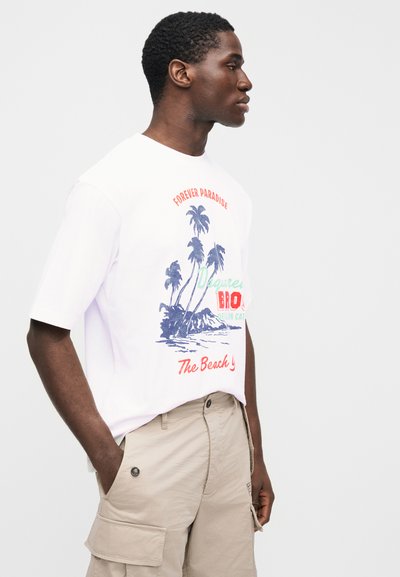 Mann in weißem T-Shirt mit Palmenmotiv und beigen Cargo-Shorts, im Profil stehend mit den Händen in den Taschen vor weißem Hintergrund.