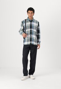 Checkered flannel shirt in green, blue, and white; button-up design with long sleeves. Paired with black trousers and white sneakers.
