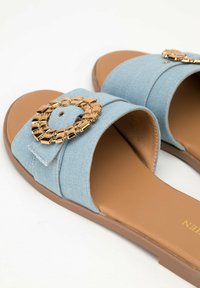 Sandali slide in denim azzurro chiaro con una fascia larga, accentuazione circolare dorata e una suola piana marrone. Materiale testurizzato con cuciture dettagliate.