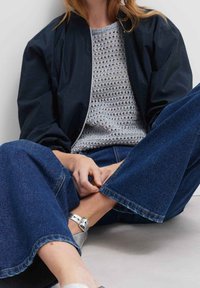 Personne assise, jambes croisées, portant une veste noire, un pull crochet gris, un jean bleu à jambes larges et des chaussures argentées avec bride à la cheville.
