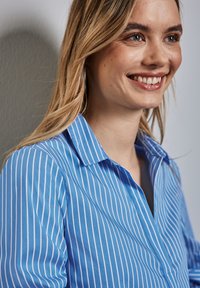 Camicia button-up azzurro chiaro con sottili righe bianche, dotata di colletto classico e tessuto testurizzato. Cuciture visibili e dettagli di design senza cuciture.