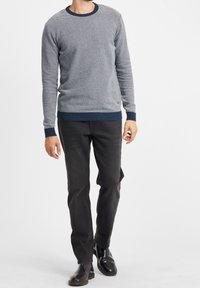 Grå texturerad stickad tröja med rund hals och marinblå ribbade ärmmuddar, kombinerad med svarta jeans och glänsande svarta skor. Mannen står avslappnat.