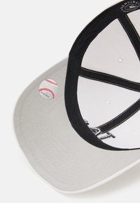 Gorra de béisbol gris con un lado negro en la parte inferior, cuenta con una pequeña etiqueta gráfica de béisbol y costuras a lo largo de la visera. El interior es blanco con detalles del logo.
