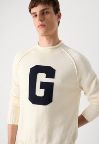 Cream knitted sweater with a ribbed collar and cuffs, featuring a large navy blue "G" logo on the chest. Rounded hem, casual fit.