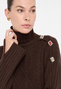 Maglione a collo alto in maglia retrò marrone, con dettagli decorativi sulle spalle in gemme rosse e trasparenti, polsini a costine e tessuto testurizzato.