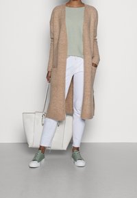 Long gilet beige, haut vert texturé, pantalon blanc et baskets vert clair. Sac cabas blanc avec design simple, ferrures visibles et accents.