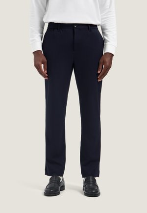 Man wearing black dress pants, black loafers, white socks, and a white long-sleeve shirt standing on a light background.