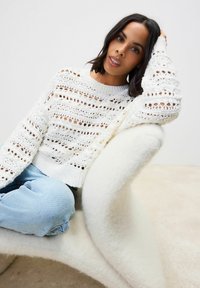 Pull en tricot blanc avec un motif ajouré, coupe décontractée, col rond, associé à un jean bleu clair. Assis sur une chaise blanche douce et courbée.