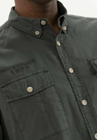 Chemise à boutons vert foncé en tissu texturé, dotée de deux poches poitrine, boutons beiges et un détail brodé "RECONNECT 1977".