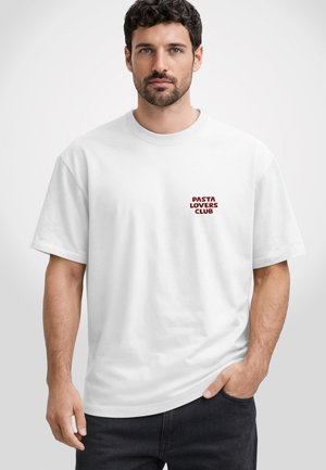 Mann mit dunklem Haar und Bart trägt ein weißes T-Shirt mit rotem Schriftzug „PASTA LOVERS CLUB“ auf der linken Brust, Hand in der Tasche.