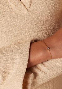 Pulsera de cadena de oro con una gema clara central, llevada en una muñeca. El fondo muestra una tela texturizada de color beige claro.
