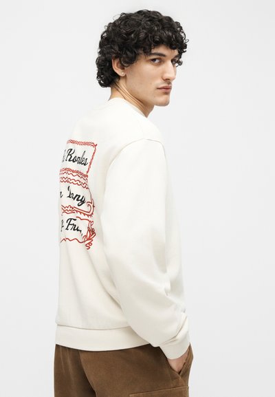 Νέος άνδρας με σγουρά μαλλιά που φοράει λευκή μπλούζα με κόκκινο σχέδιο γραφής στην πλάτη, καφέ παντελόνι και το χέρι στην τσέπη, κοιτάζοντας πάνω από τον ώμο.