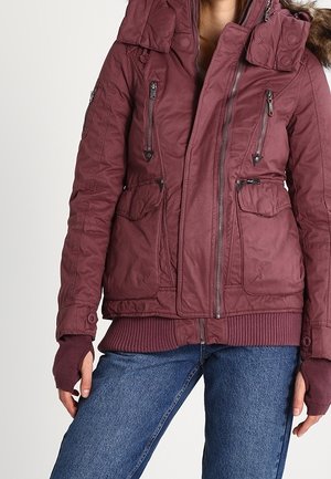 Osoba w bordowej zimowej kurtce z wieloma kieszeniami na zamek i rękawiczkami bez palców, w połączeniu z niebieskimi dżinsami.