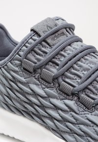 Grå textilsneakers med en vävd ovandel, grå snören och en mockapatch på tungan. Har en vit gummisul och en strukturerad design.