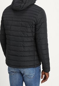 Svart puffjacka med horisontellt quiltat mönster, med en huva och en slät yta. Bärs med blå jeans.