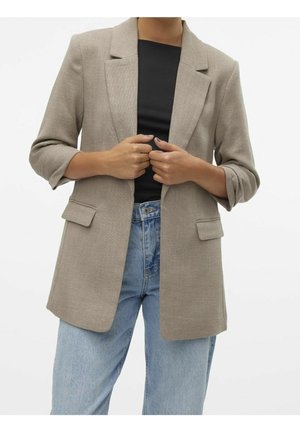 Persoon die een beige blazer met opgestroopte mouwen aanpast, draagt een zwarte top en lichtblauwe jeans tegen een effen witte achtergrond.