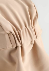 Bež zbrano blago z gladkim teksturo in rahlo raztegnjenimi podrobnostmi. Vsebuje subtilno šivanje vzdolž zbrane šivnice.