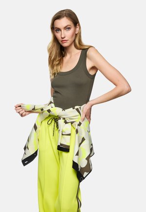 Sportoutfit mit einem olivgrünen Tanktop und hellgelben, strukturierten Hosen, dazu ein gemusterter Wickel in neutralen Farbtönen, der auf der Taille gebunden ist.