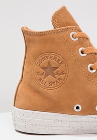 Tan leather high-top sneaker with a textured finish, featuring a circular logo, white speckled sole, and metal eyelets for laces.