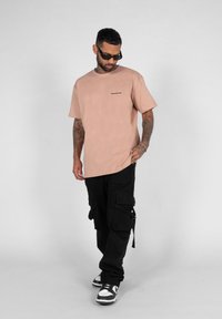 Lys rosa oversized t-skjorte med et trykt logo, kombinert med svarte cargo-bukser med flere lommer og stropper, iført svarte joggesko.