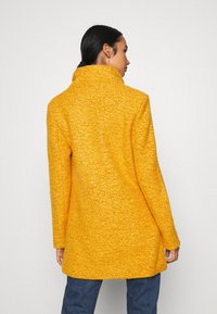 Cappotto giallo con texture, colletto alto, maniche lunghe e taglio dritto, abbinato a jeans blu. La vista posteriore mette in evidenza la lunghezza del cappotto.