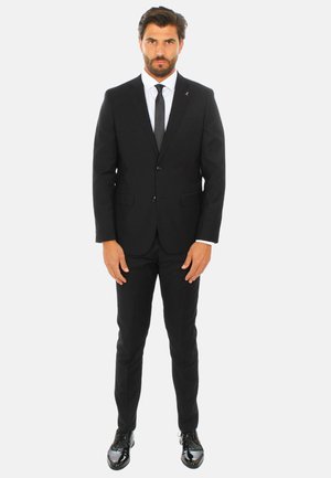 Completo nero con un design aderente, dotato di giacca monopetto, camicia bianca e cravatta nera. Scarpe lucide nere completano il look.