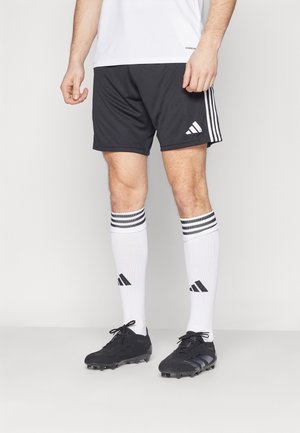 Mężczyzna w czarnych szortach Adidas, białych, sięgających do kolan skarpetach Adidas z paskami oraz czarnych korkach piłkarskich na jednolitym tle.