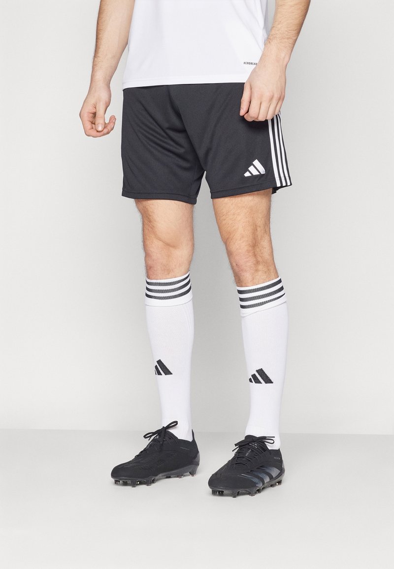 Homme portant un short noir Adidas, des chaussettes blanches Adidas montantes à rayures et des crampons de football noirs sur un fond uni.