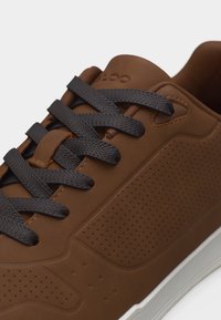 Baskets marron avec des lacets noirs, des panneaux latéraux perforés et une semelle blanche, montrées en détail rapproché.