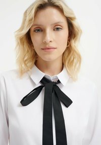 Blusa blanca con cuello puntiagudo y una pajarita negra. Textura de tejido suave con largas cintas negras que caen.