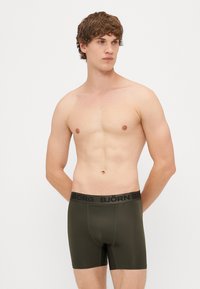 Olīvu zaļi bokseri ar melnu zīmola elastīgu jostu, izgatavoti no gludas auduma ar piegulīgu dizainu. Nav redzamu rakstu vai akcentu.