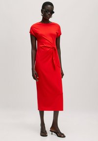 Rotes, knielanges Kleid mit kurzen Ärmeln und seitlichem Bindeband aus glattem Stoff. Das Model trägt schwarze Sonnenbrillen und Sandalen.
