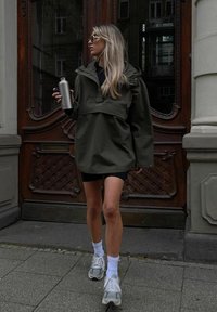 Femme aux longs cheveux blonds portant des lunettes de soleil, une veste verte oversize, un short noir, des chaussettes blanches et des baskets, tenant une bouteille argentée près d'une porte en bois.