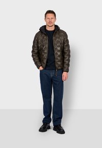 Quilted brown padded jacket with a black hood, paired with a black sweater and blue jeans. Model stands with hands in pockets on a plain background.