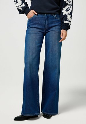 Persoon die een high-waisted blauwe wijde jeans draagt, zwarte puntige schoenen en een donkere trui met witte bloemenpatronen op de mouwen.