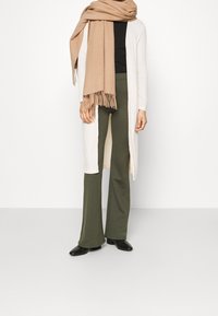 Larga y ligera chaqueta de punto color crema, top negro, pantalones de campana verde oliva y una bufanda beige con flecos. Zapatos negros de punta afilada.